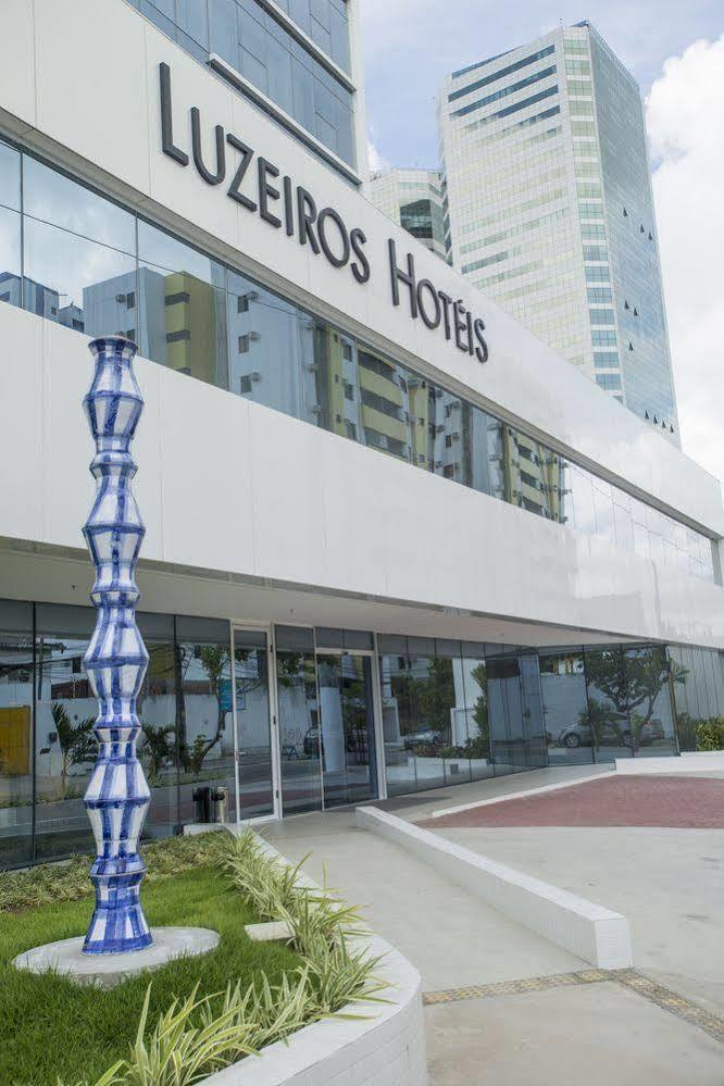 Hotel Luzeiros Ресифи Экстерьер фото The photo shows a modern hotel building with large glass windows and a sleek, white facade. In front of the hotel, there is a tall, decorative sculpture that features a unique, twisting shape with a blue and white color scheme. The hotel name is prom